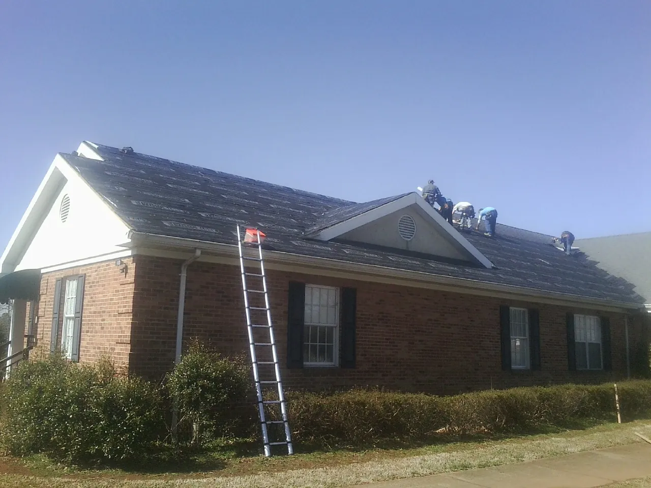 Crew tearing shingles off a brick ranch roof with a ladder set — Emergency Roof Repair work by our crew in Birchwood