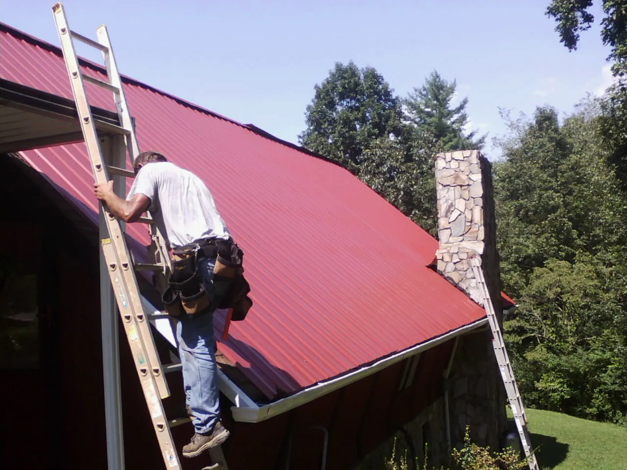 Roofer on a ladder beside a red metal roof and stone chimney — Soffit & Fascia Repair by Roofing Brothers in Birchwood